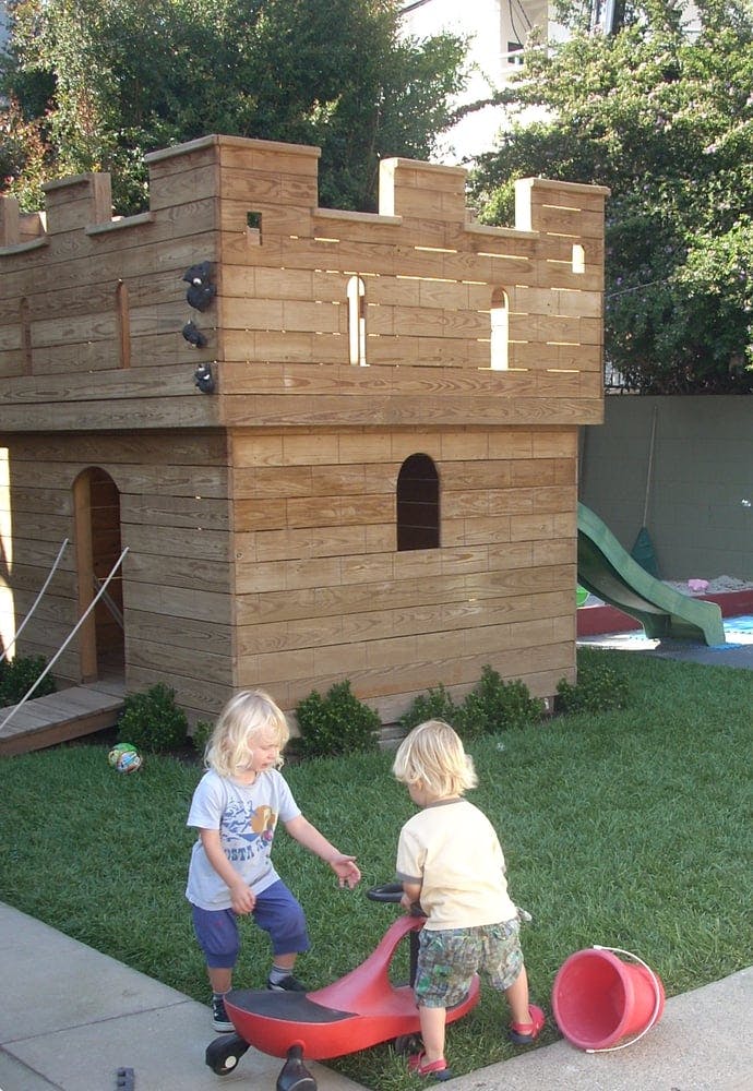 Two children playing with a red ride-on toy in a yard with a wooden playhouse