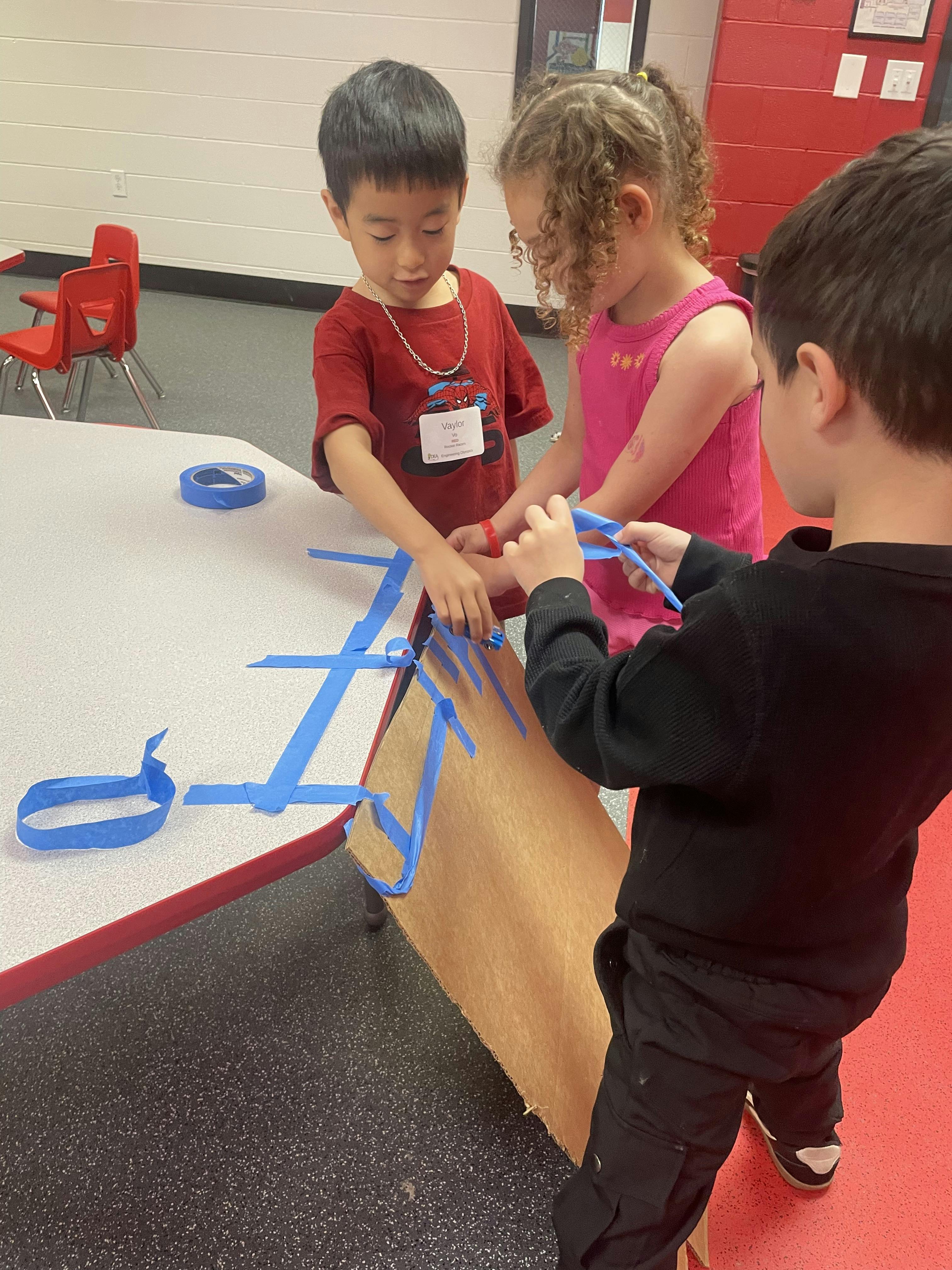 Three children taping blue strips to a piece of cardboard in a classroom