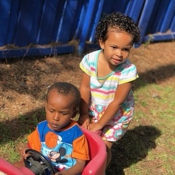 Child sitting in toy car with another child standing behind