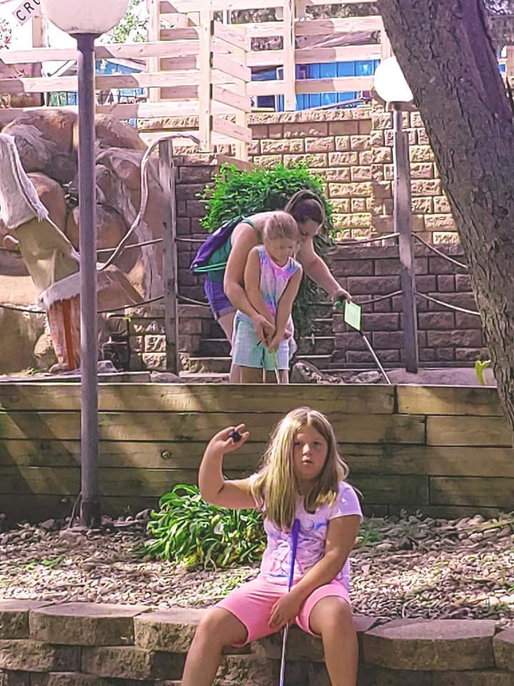 Person and child playing mini golf, another child sitting in foreground