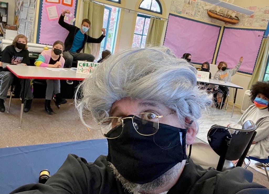 Person in wig and mask taking a selfie with students in a classroom