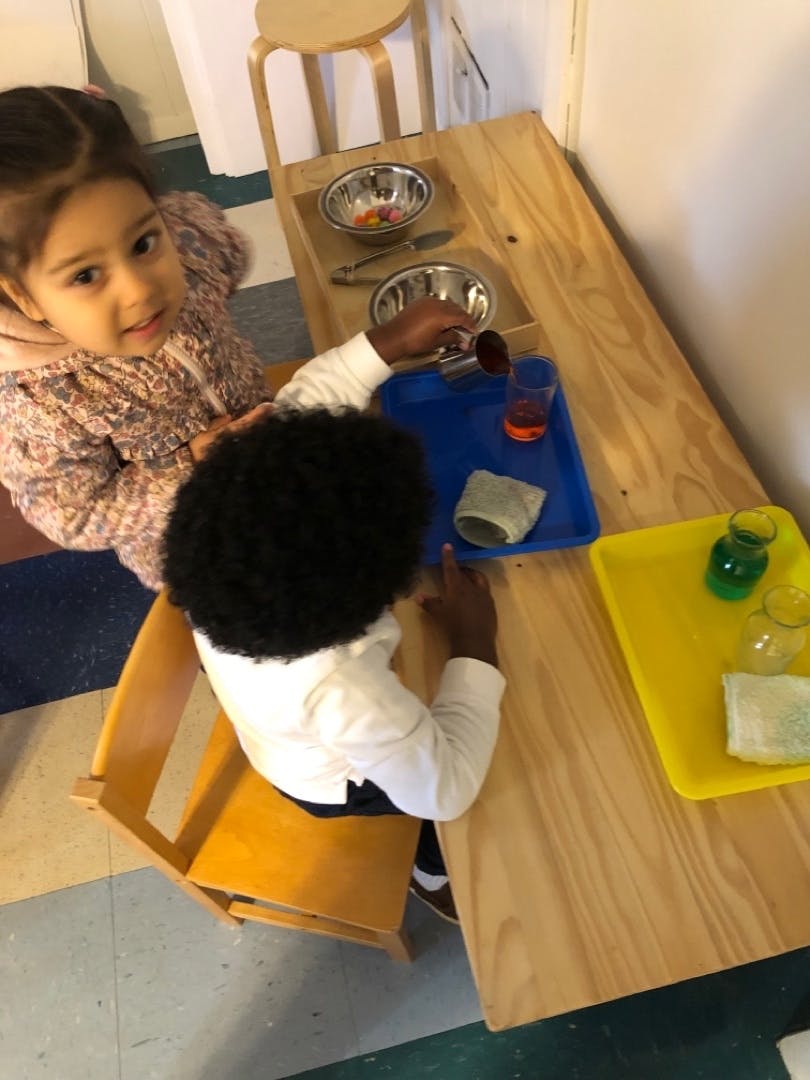 Child pouring liquid from a metal pitcher into a glass on a table