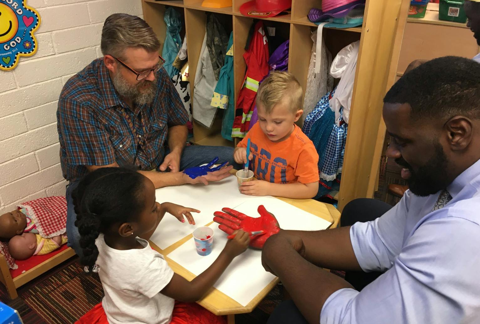 Two adults and two children at a table with one child touching a red handprint