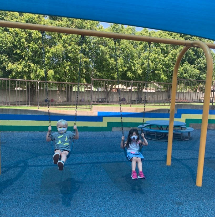 Two children wearing masks on swings at a playground