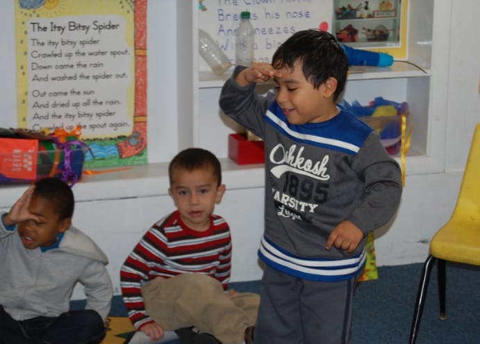 Three children in a classroom, one standing with hand to forehead