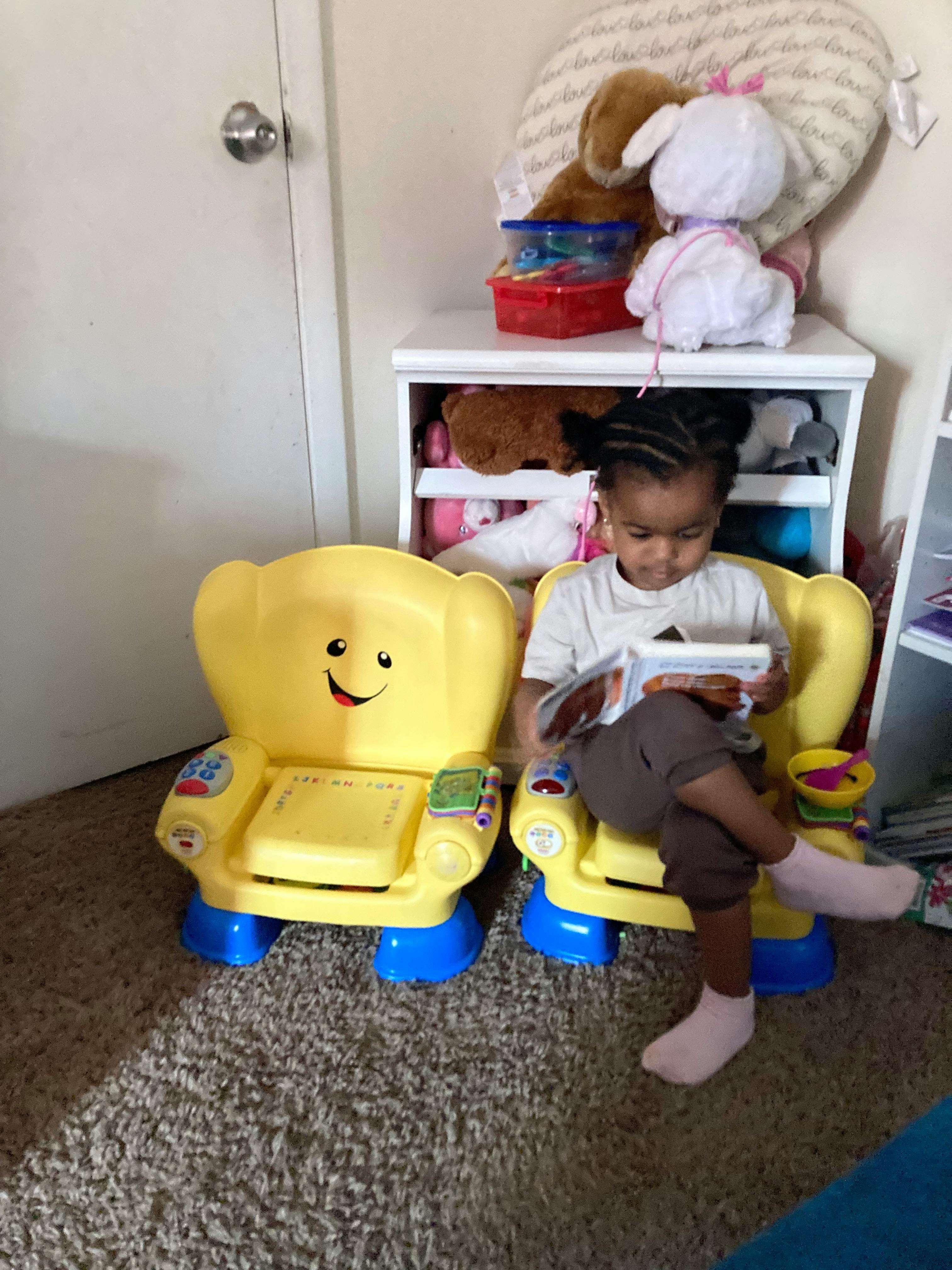 Child sitting in a yellow toy chair while reading a book
