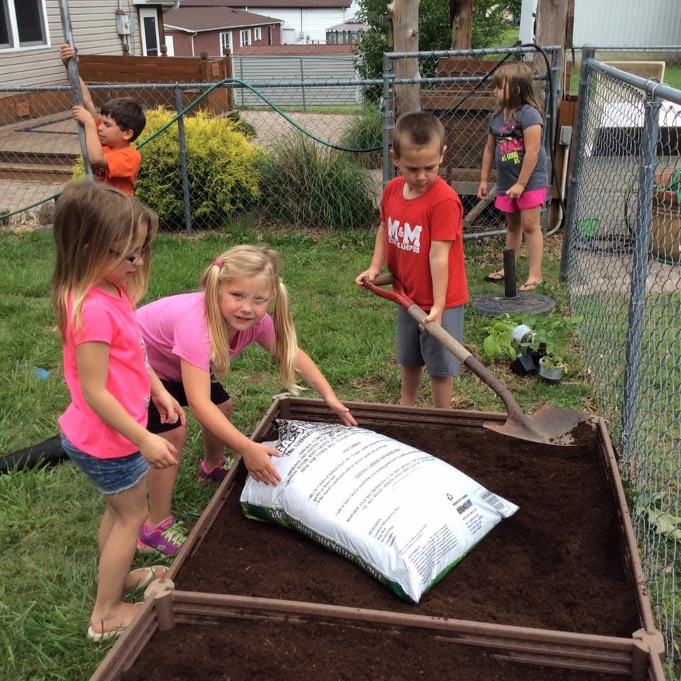 Children gardening, one holding a bag of soil, another with a shovel