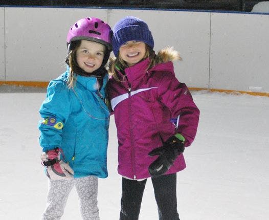 Two children in winter coats and helmets on an ice rink
