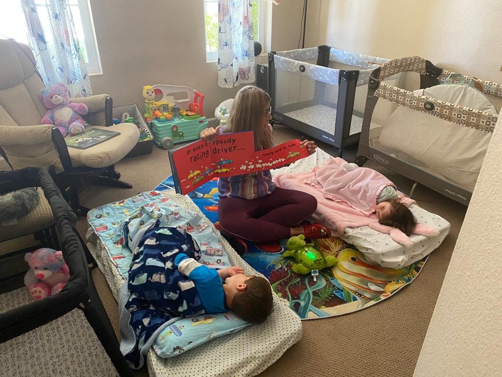 Child reading a book to two sleeping children in a room with cribs