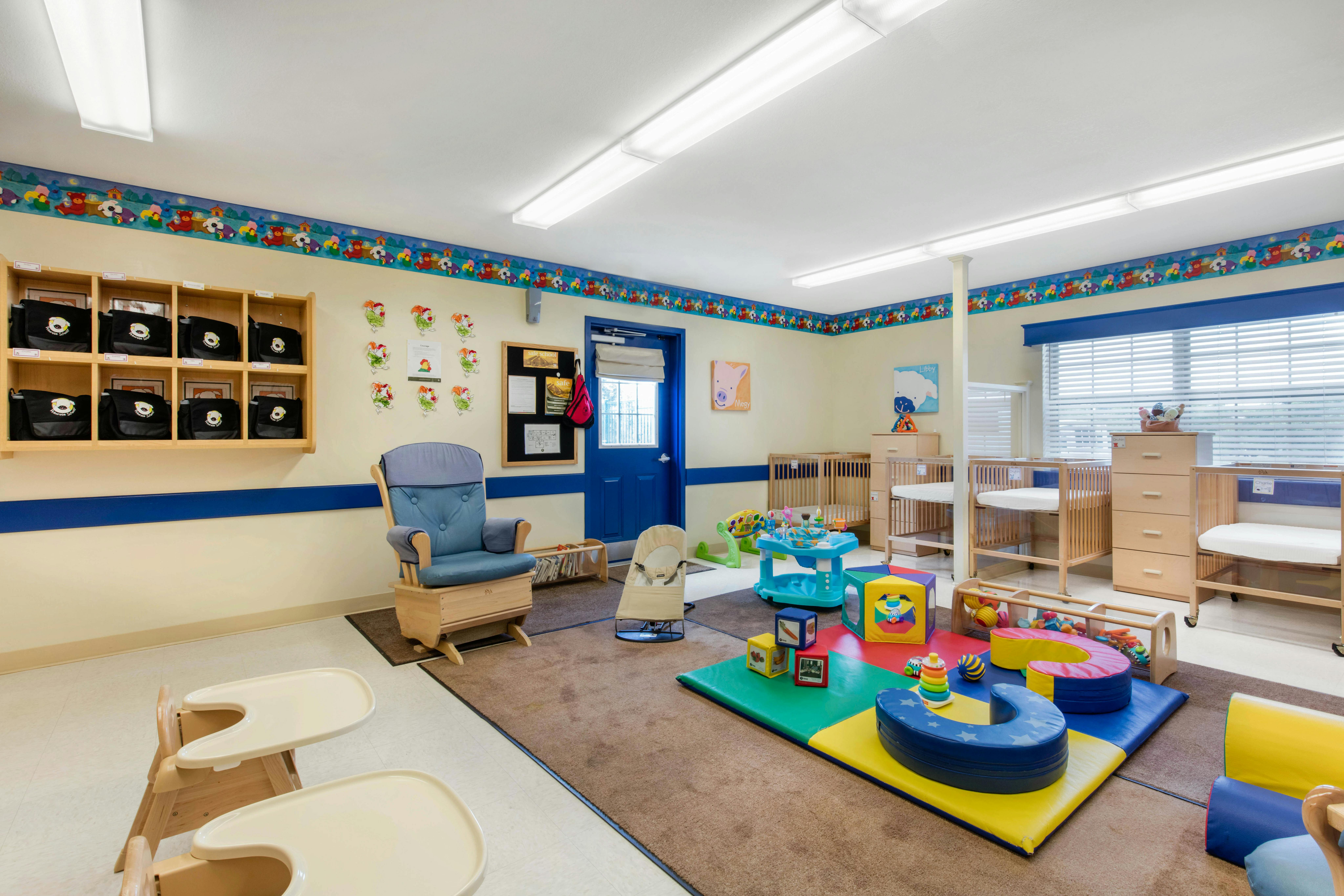 A colorful childcare room with cribs, play mats, high chairs, and a rocking chair