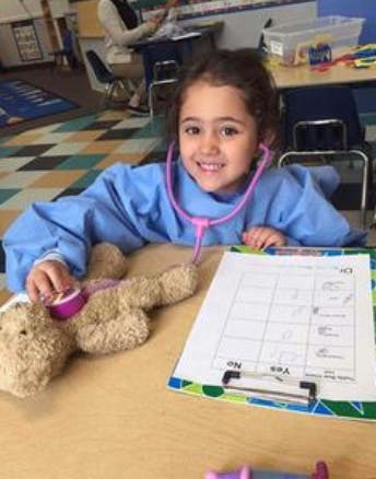 Child in blue smock with stethoscope examining a teddy bear