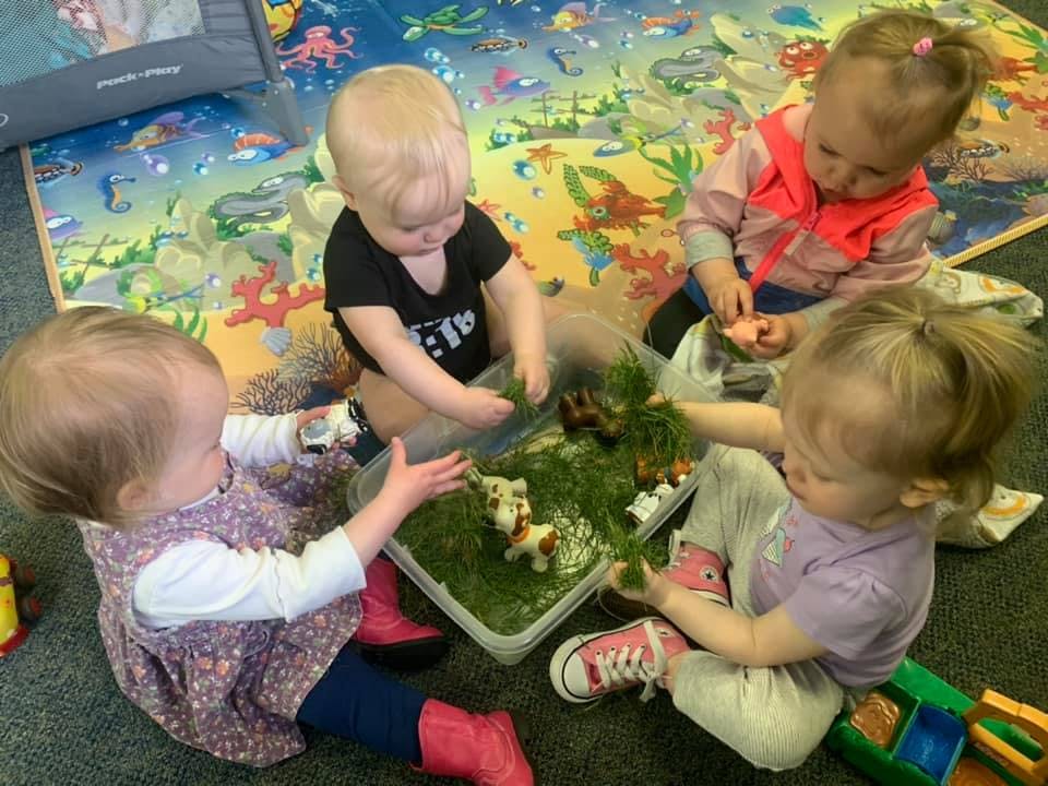Four children playing with a sensory bin filled with green material and brown items