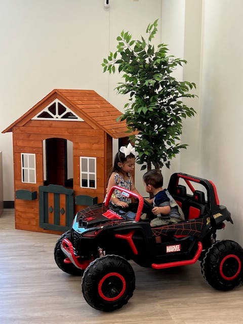 Two children in a toy car next to a playhouse