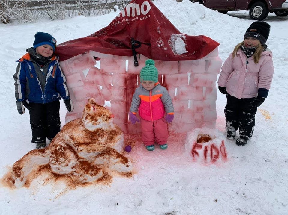 Three children standing in snow next to a snow fort and snow sculpture