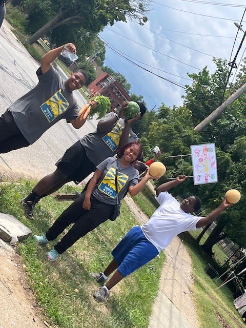 Four teens holding up various fruits and vegetables outdoors