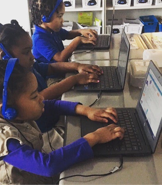 Three children wearing headphones and typing on laptops in a classroom