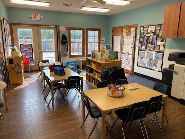 Empty classroom with small tables and chairs, exit sign above glass doors