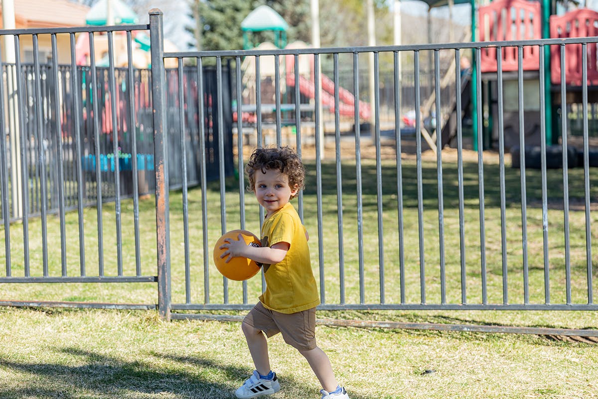 Child in yellow shirt holding a ball on grass near a fence