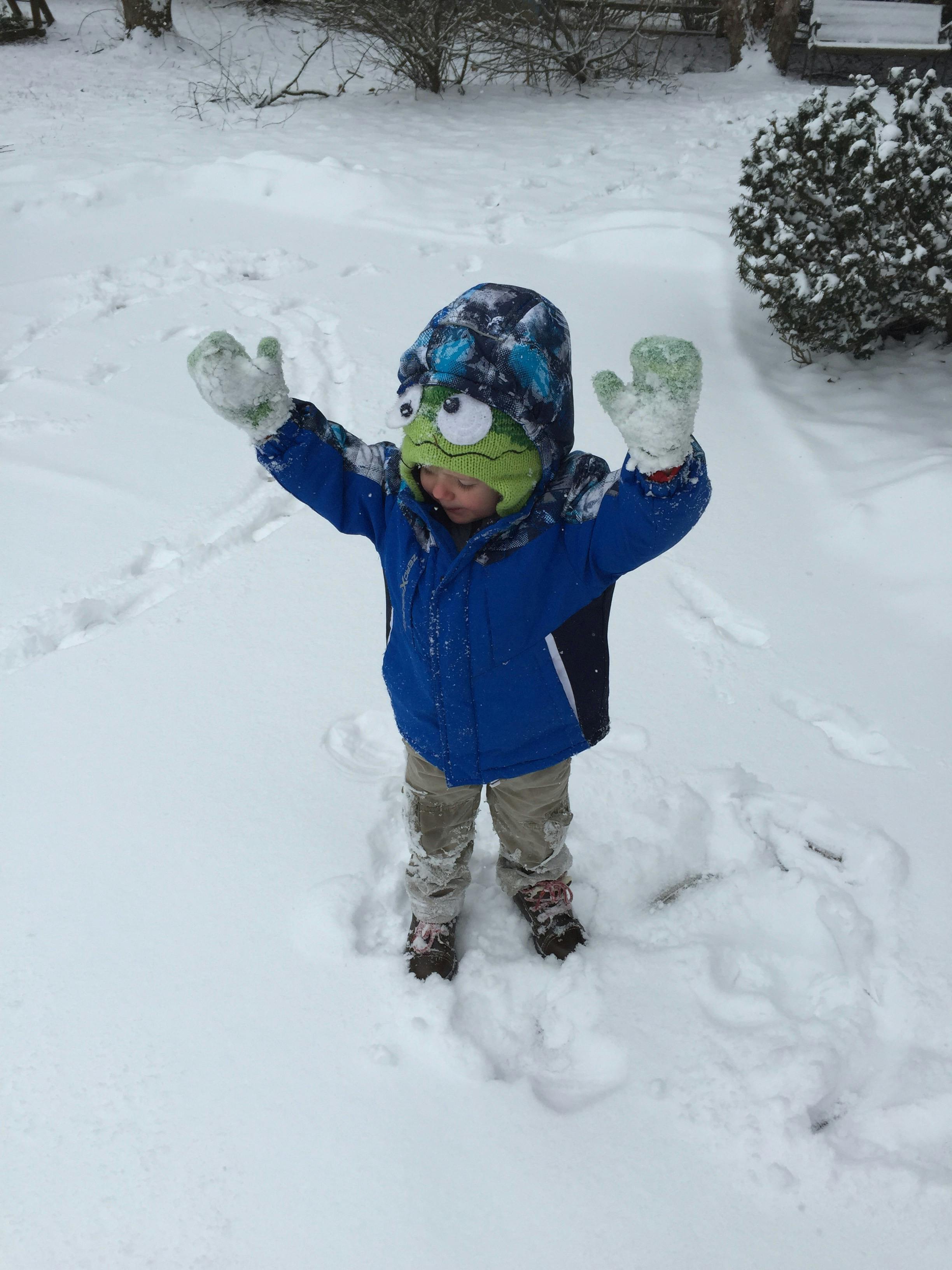 Child in a blue coat and green hat standing in snow with arms raised