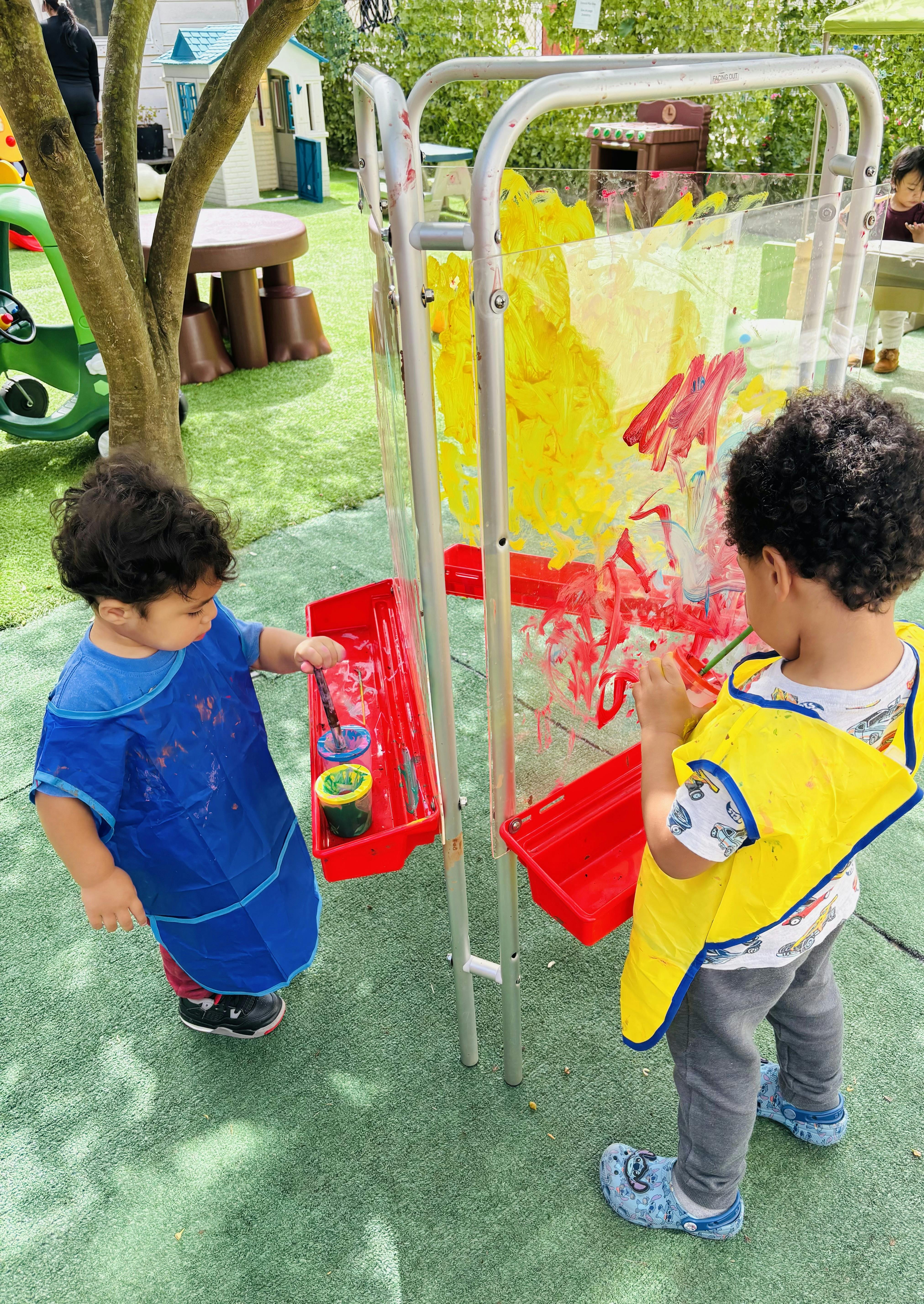Two children painting on a clear standing easel outdoors