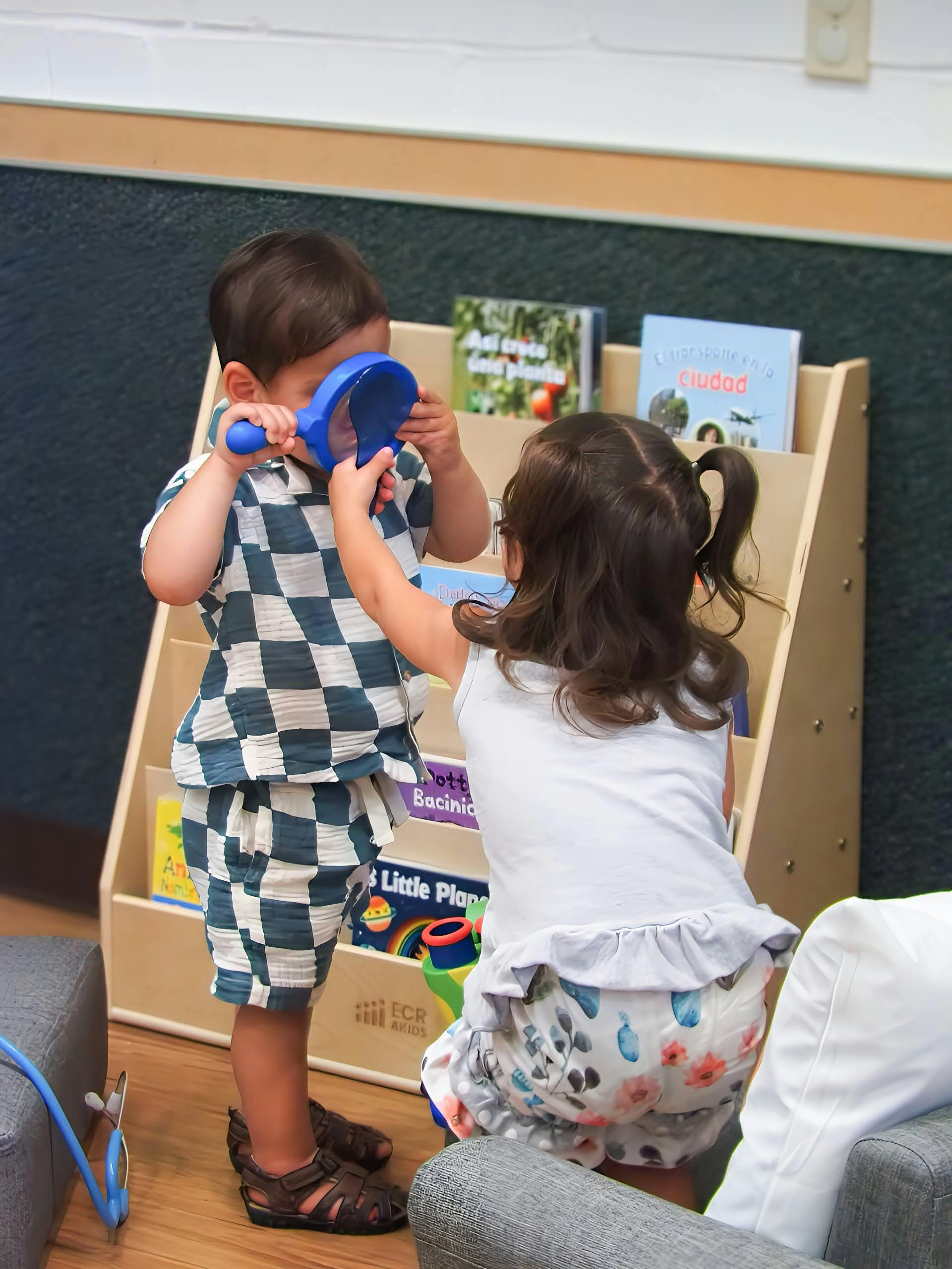 Two children playing with a blue magnifying glass near a bookshelf