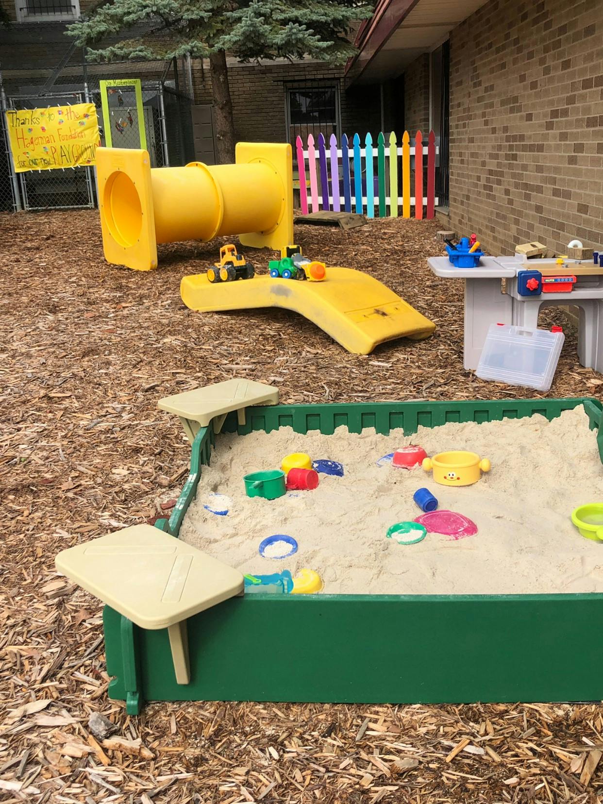 Outdoor playground with a yellow tunnel slide, sandbox, and colorful fence