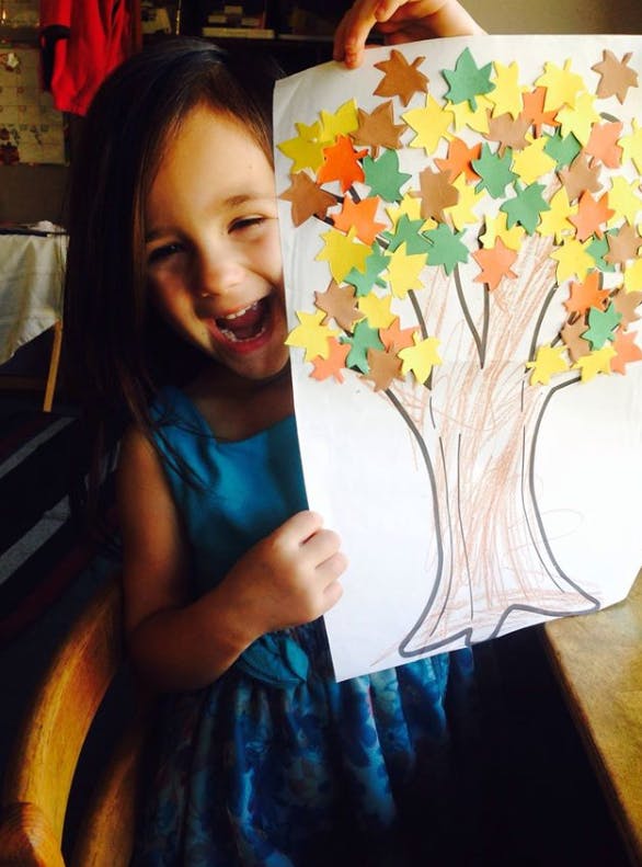 Child holding up a drawing of a tree with colorful leaf cutouts