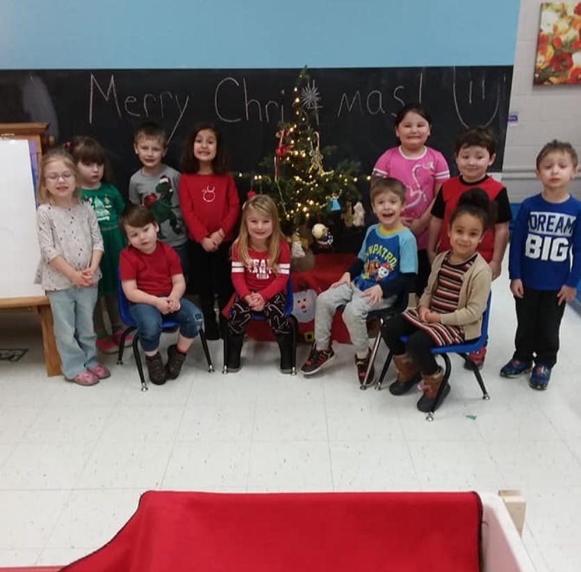 Group of children posing in front of a Christmas tree and a chalkboard with "Merry Christmas"