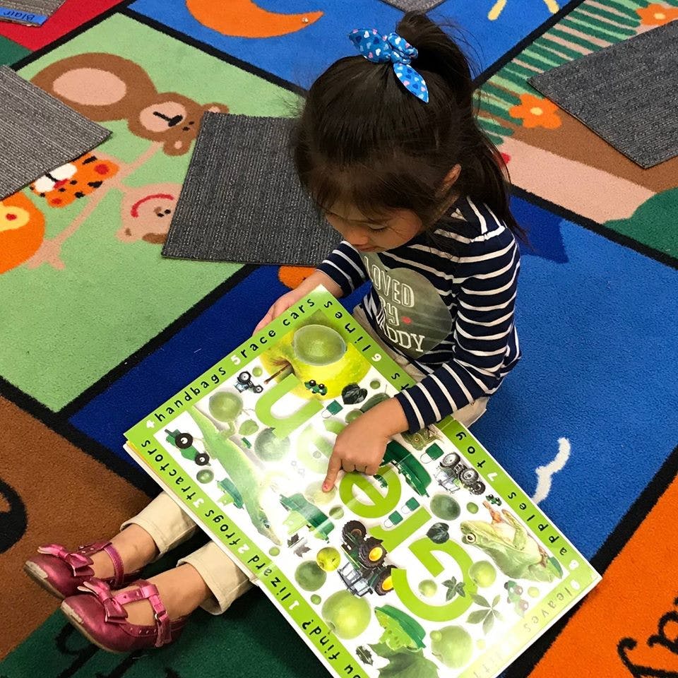 Child sitting on a colorful rug, pointing at a large green book