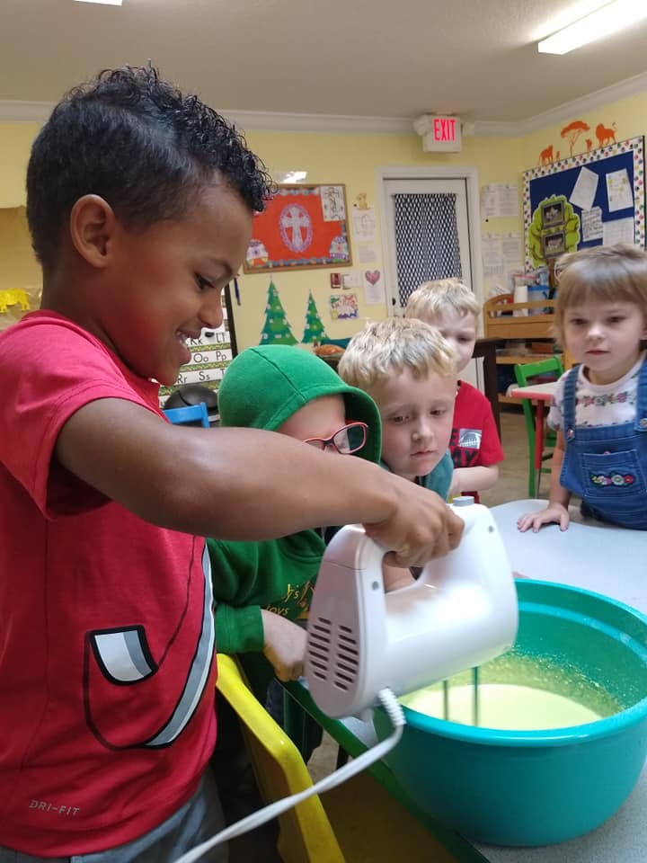 Child using a hand mixer in a bowl with other children watching