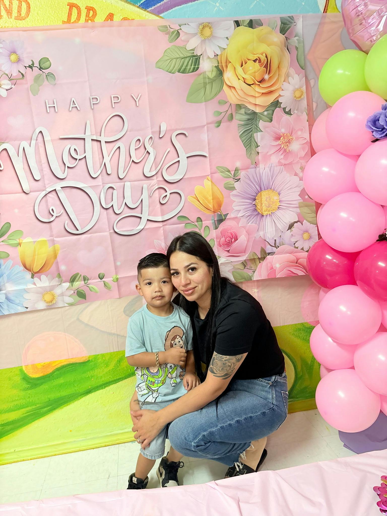 Person crouching beside a child in front of a Mother's Day backdrop