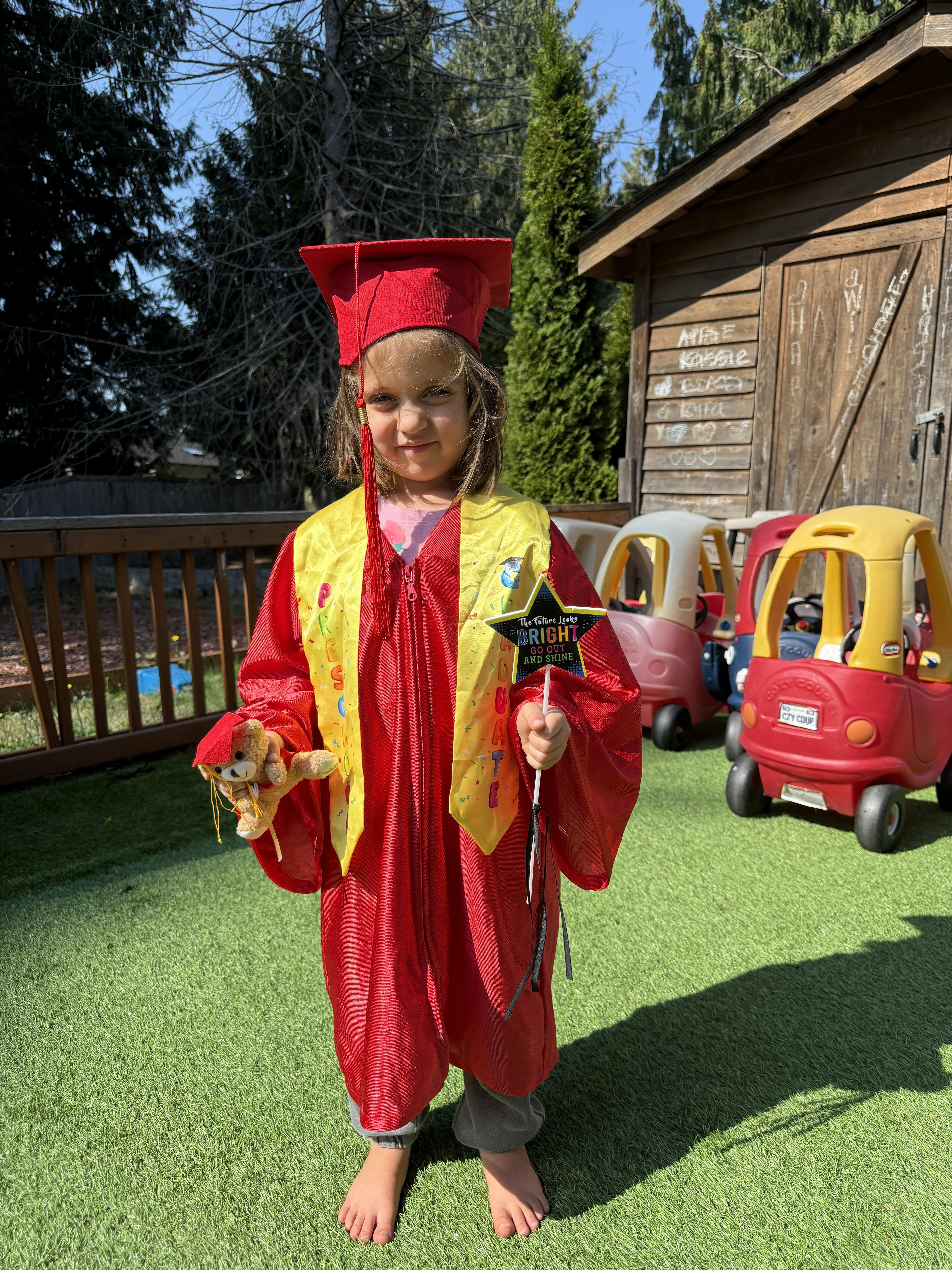 Child in red graduation gown and cap holding a star wand and stuffed animal