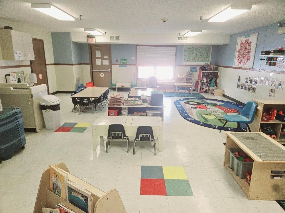 An empty classroom with small tables and chairs, a play rug, and storage shelves