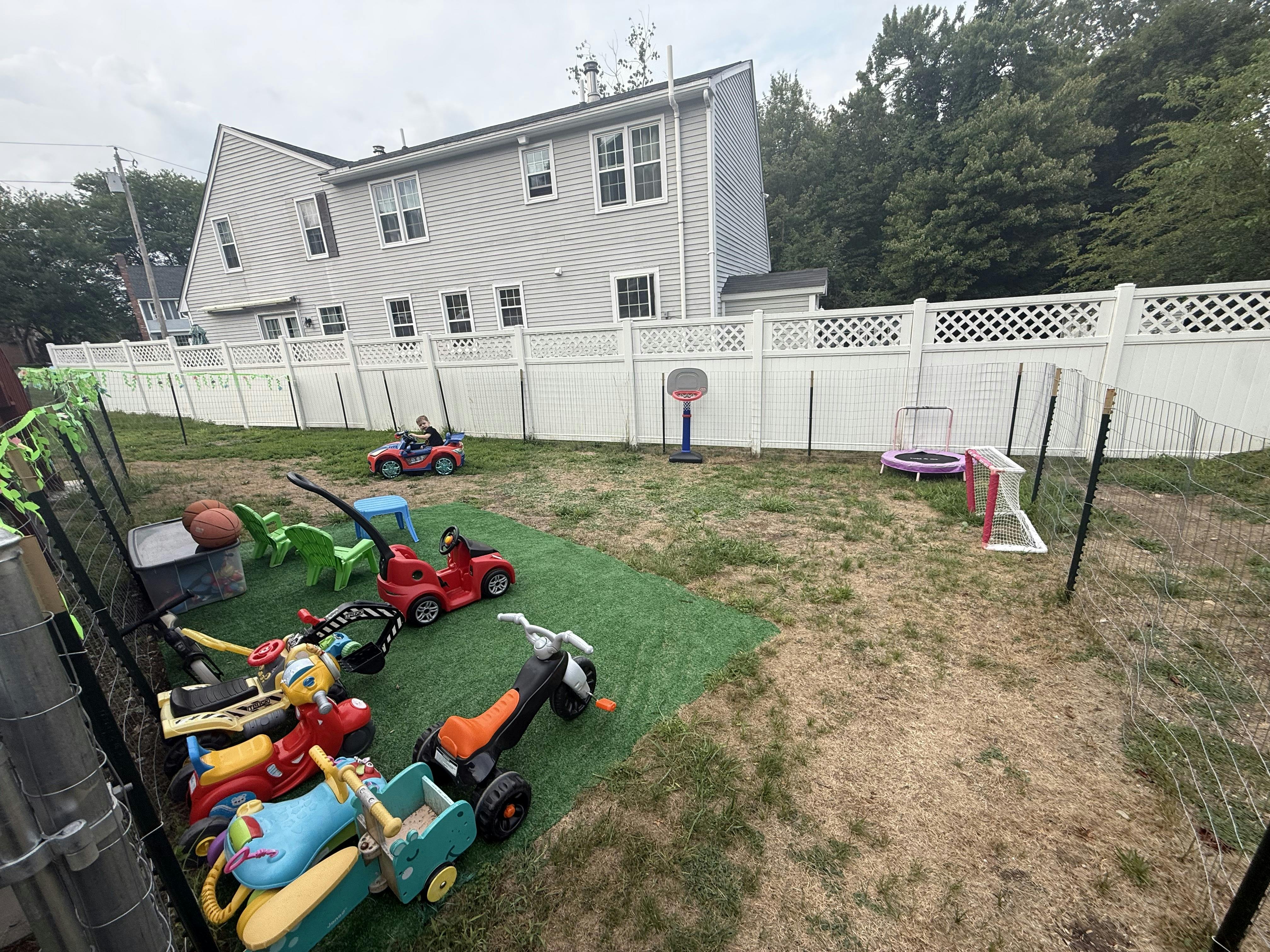Child riding a toy car in a fenced residential backyard with play equipment
