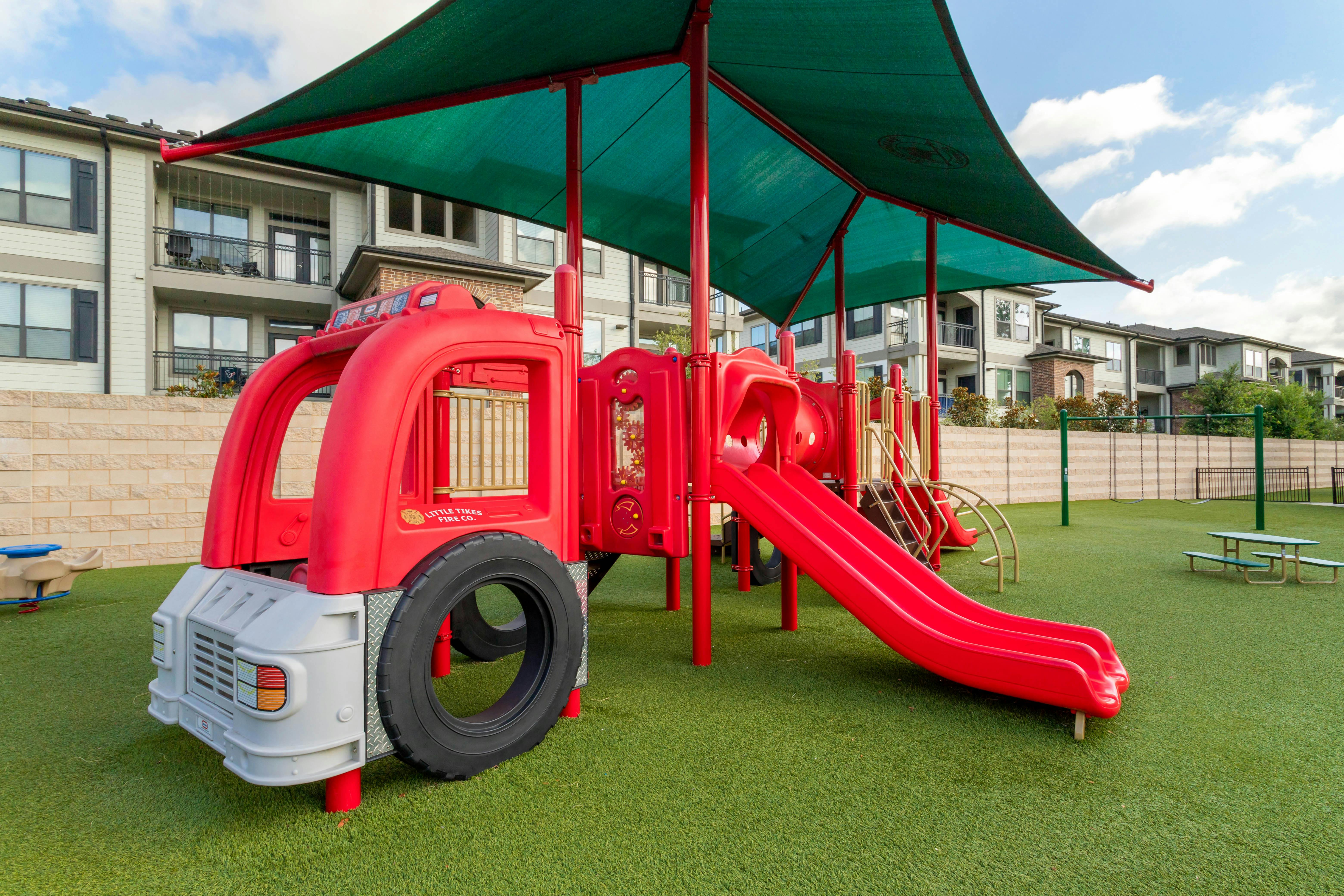 Red fire truck-shaped playground with slides and a green canopy on artificial grass