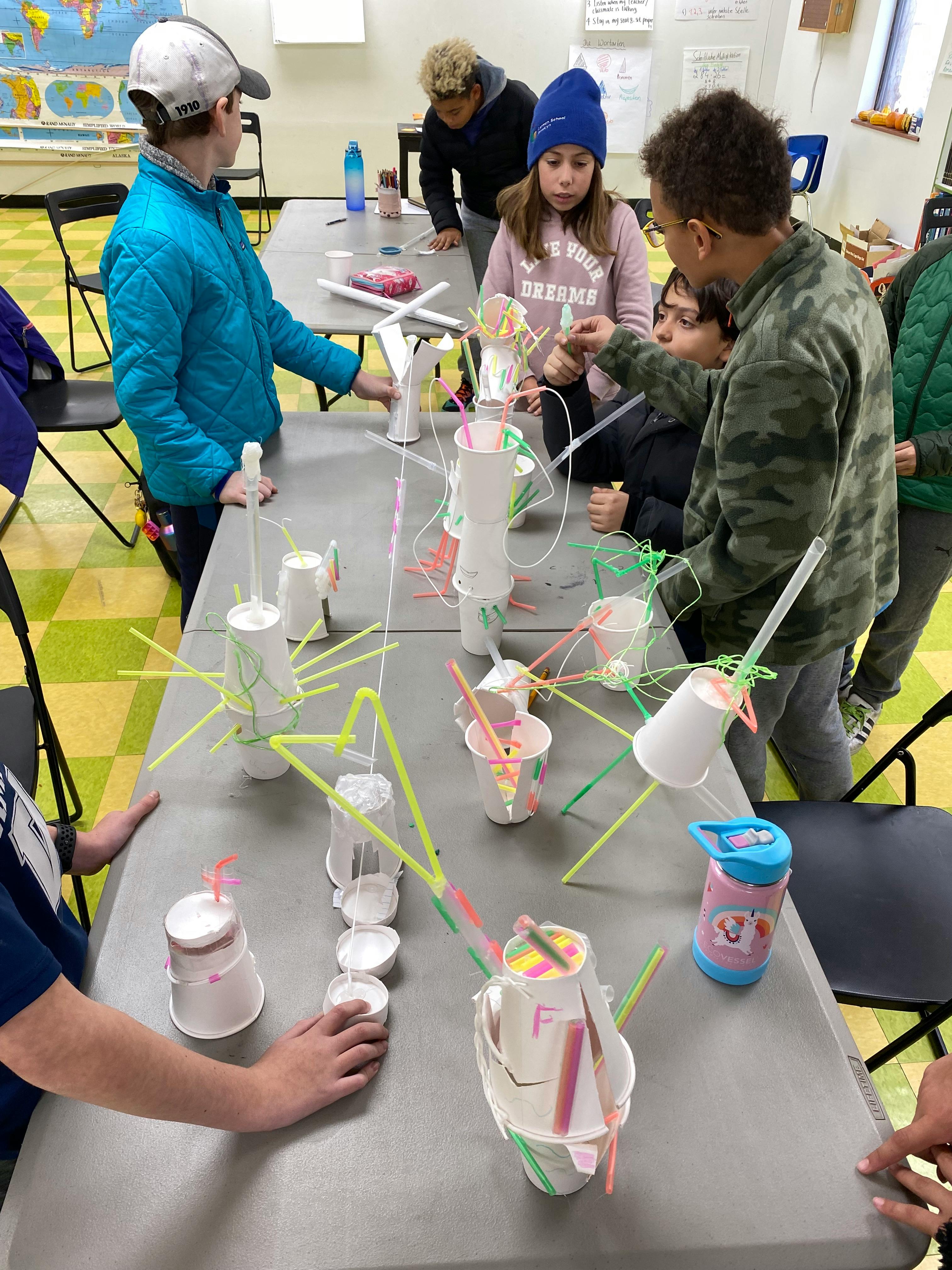 People building structures with cups and straws on a table in a classroom