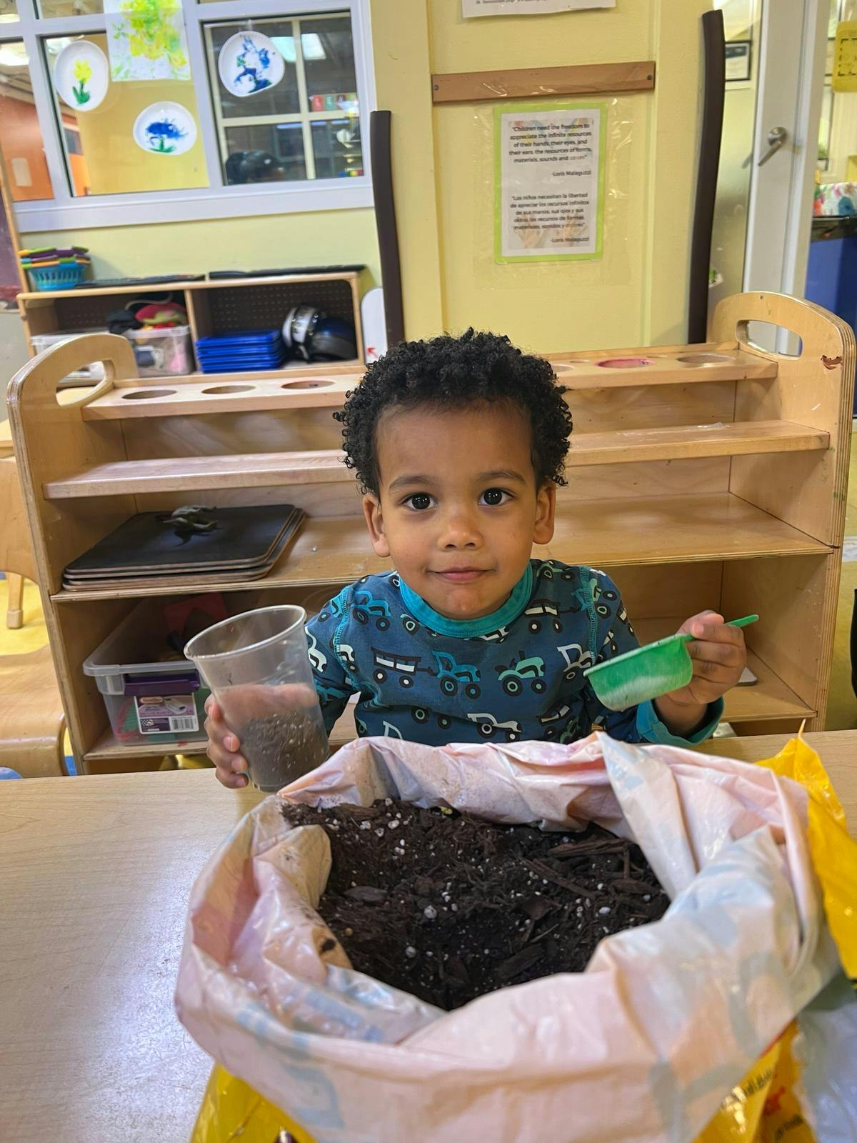 Child holding a cup of soil and a small green scoop next to a bag of soil