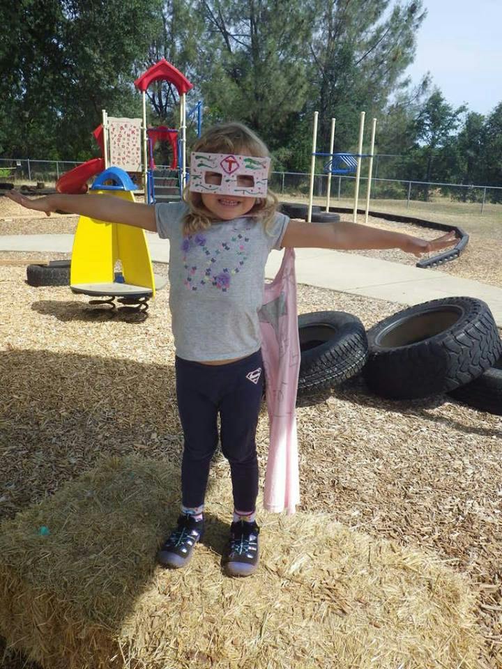 Child wearing a mask and cape with arms outstretched on a playground
