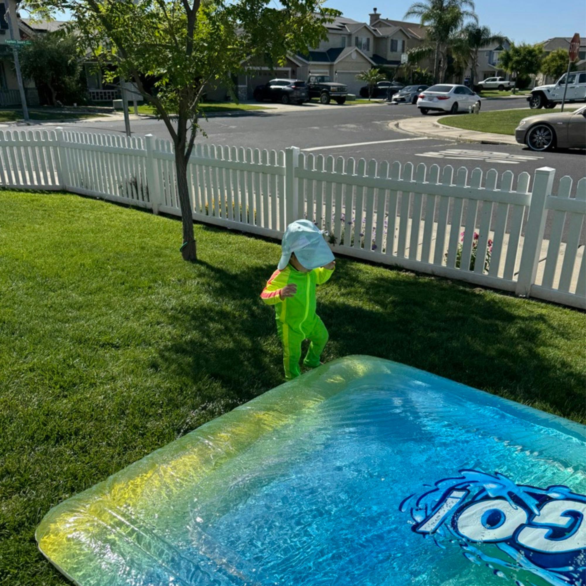 Child in a green suit and blue hat next to a water play mat on grass