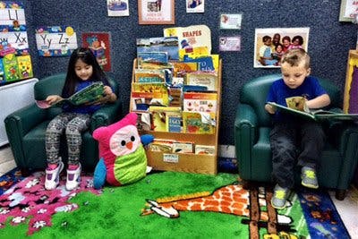 Two children sitting in chairs reading books in a classroom