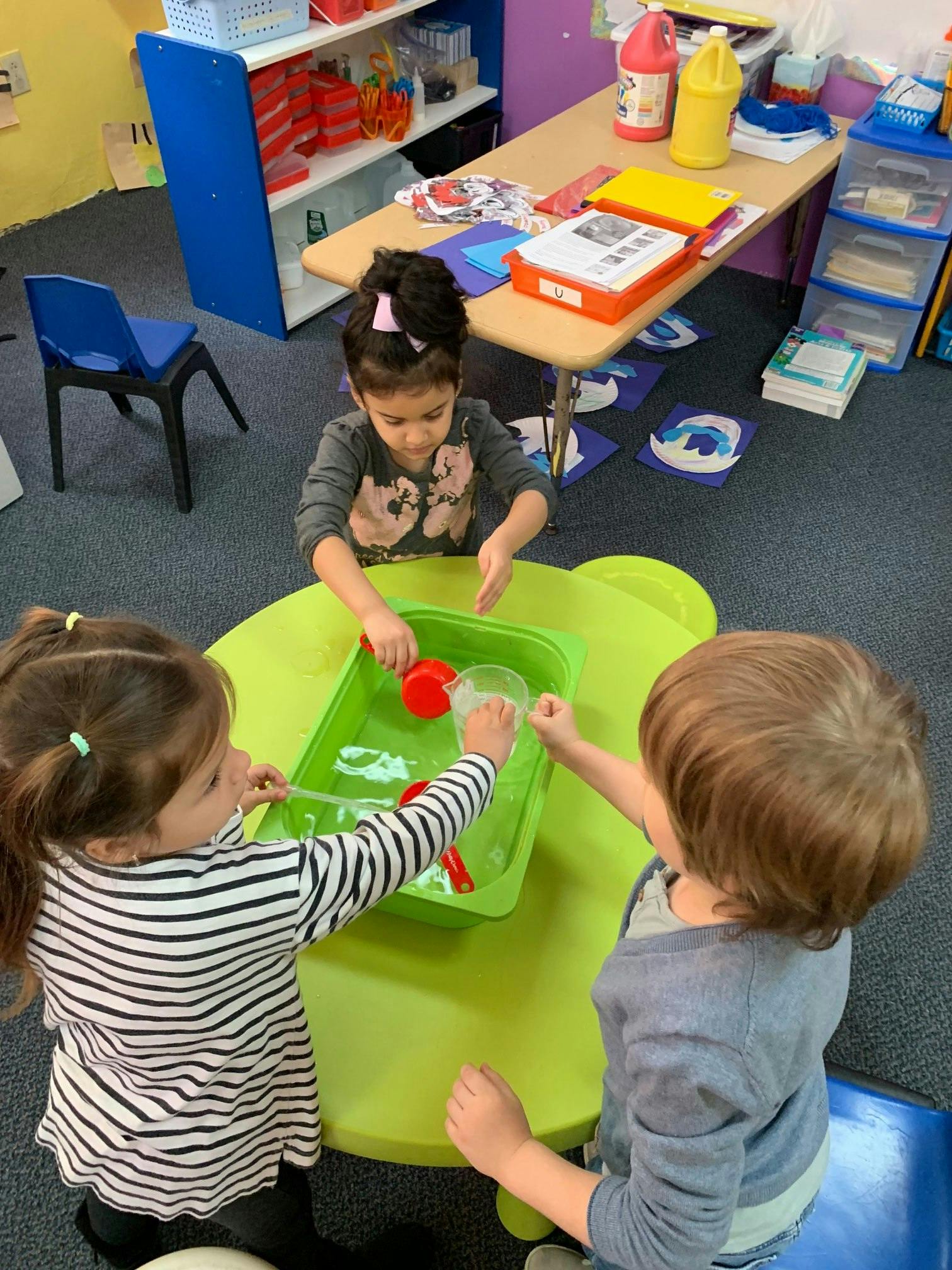 Three children playing with water in a green bin in a classroom