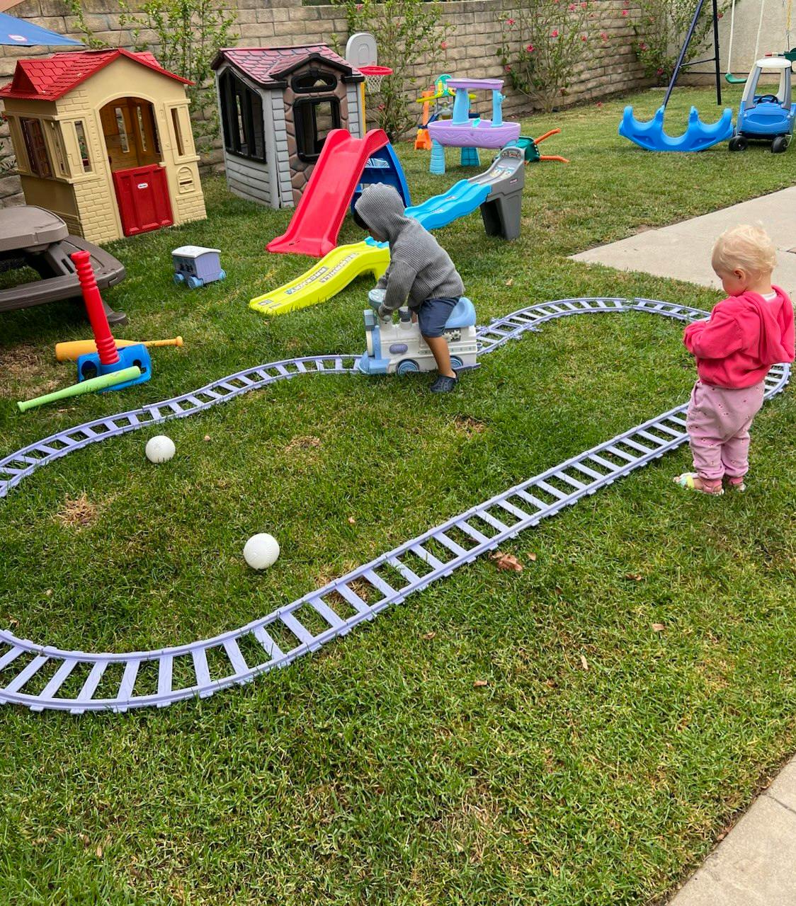 Child playing with a toy train on tracks in a grassy yard with playhouses