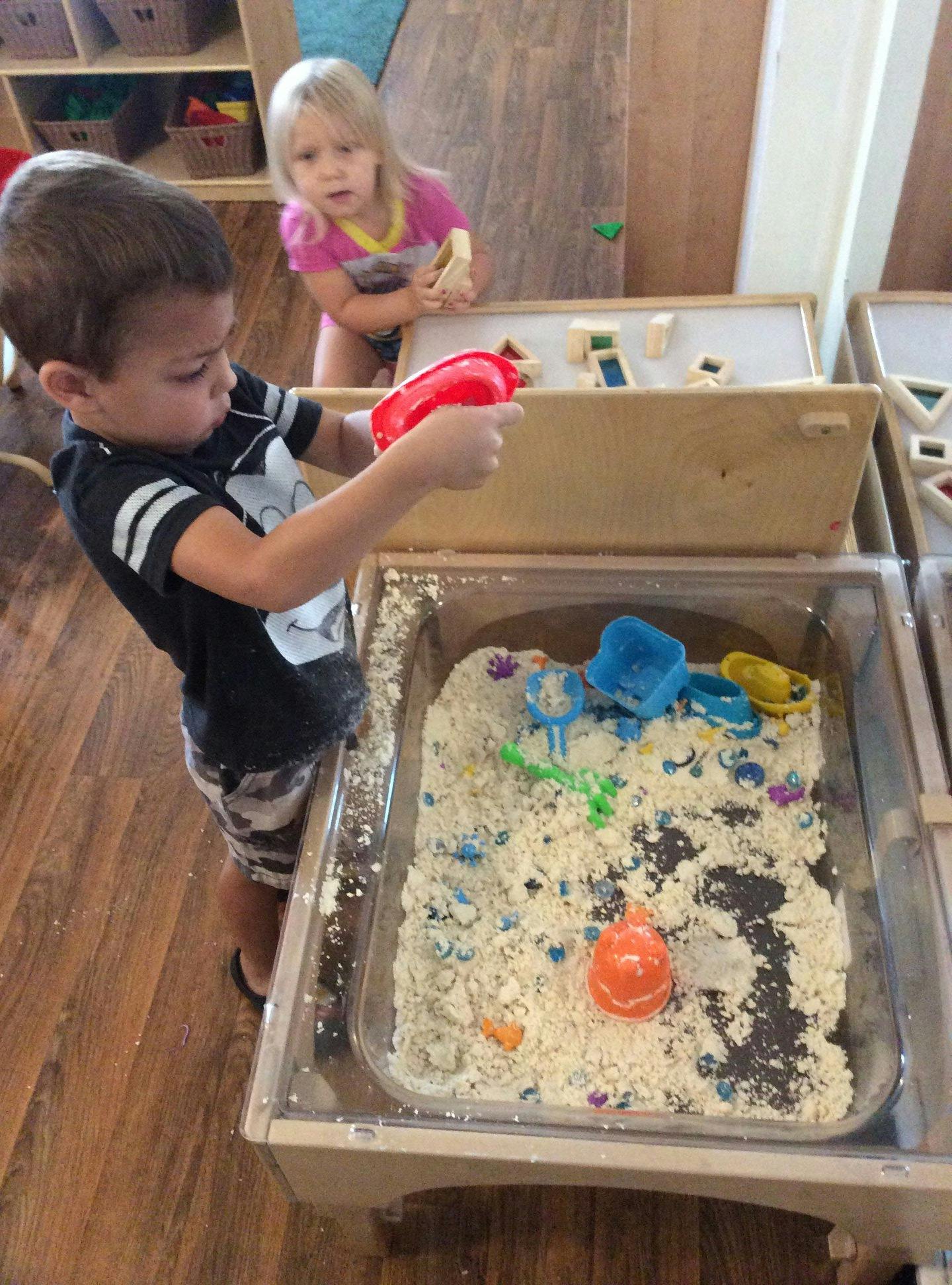 Child playing with a red scoop in a sand table, another child in background