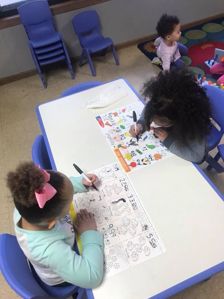 Two children drawing at a table in a classroom, another child on the floor