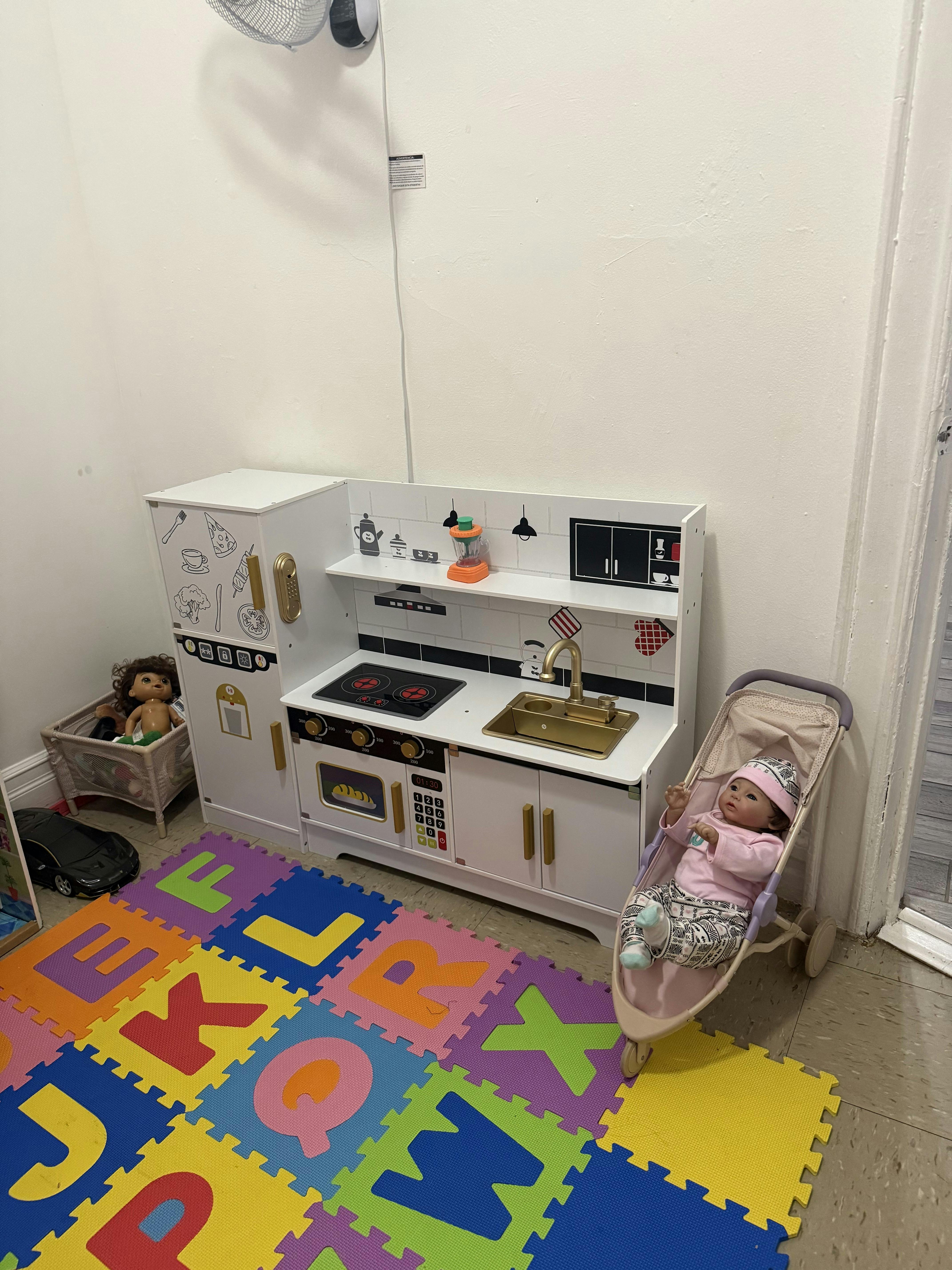 Doll in a stroller next to a toy kitchen set and alphabet floor mats
