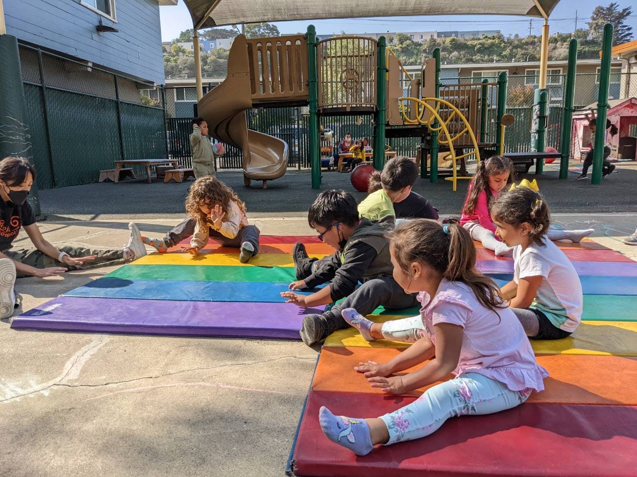 Children and an adult sitting on rainbow mats on a playground