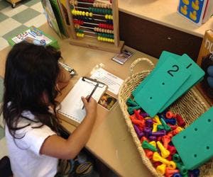Child writing on a clipboard at a table with a basket of colorful objects