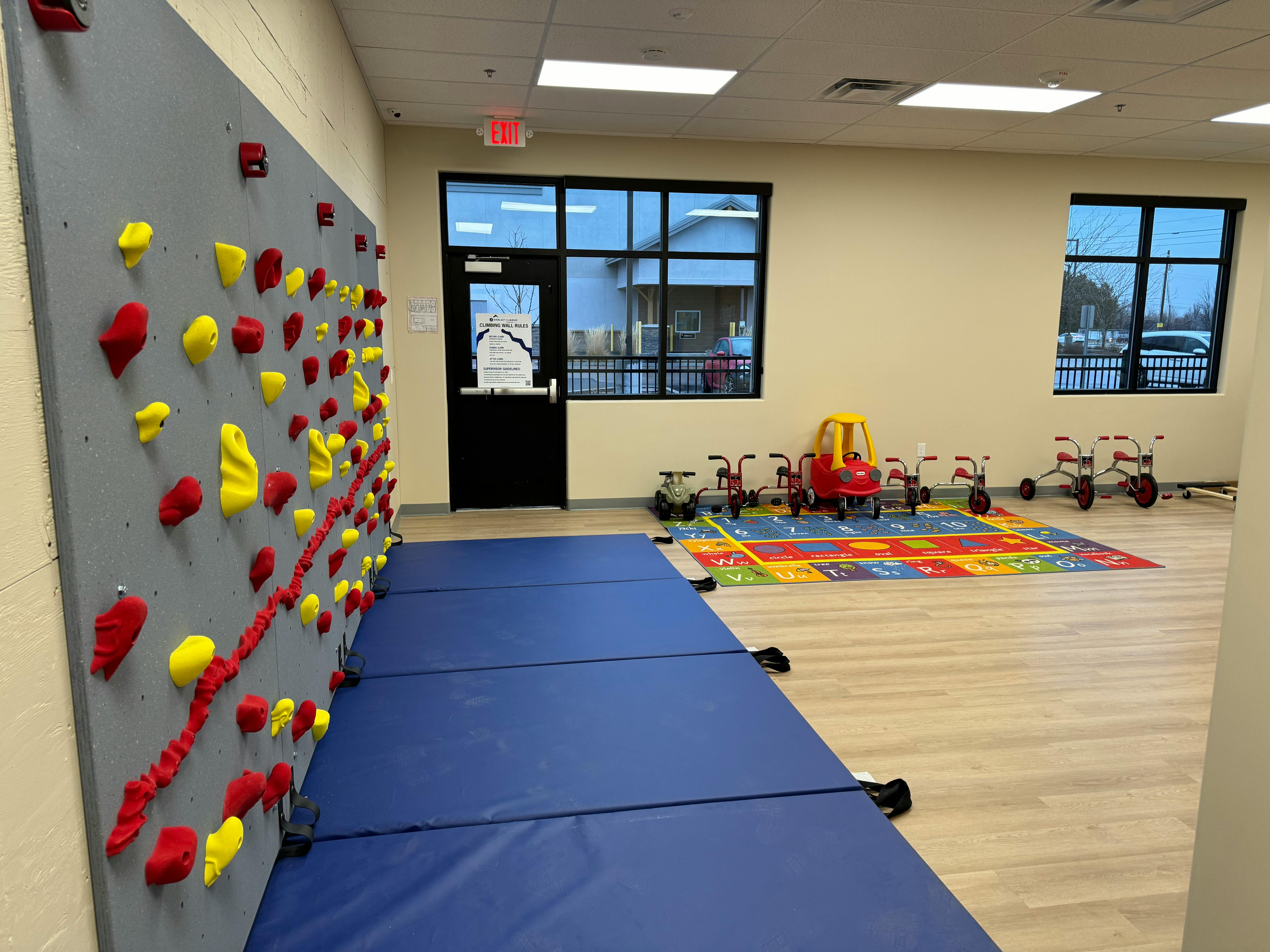 Indoor climbing wall with blue mats in a room with small tricycles