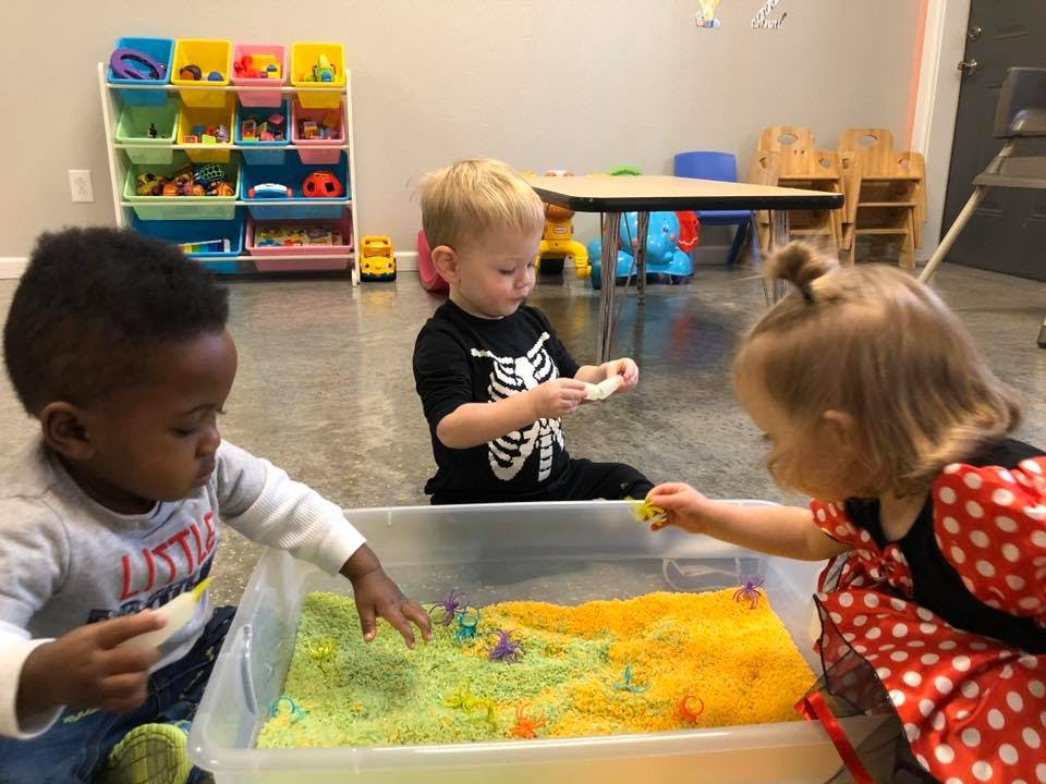 Three children playing with colorful sand in a clear bin