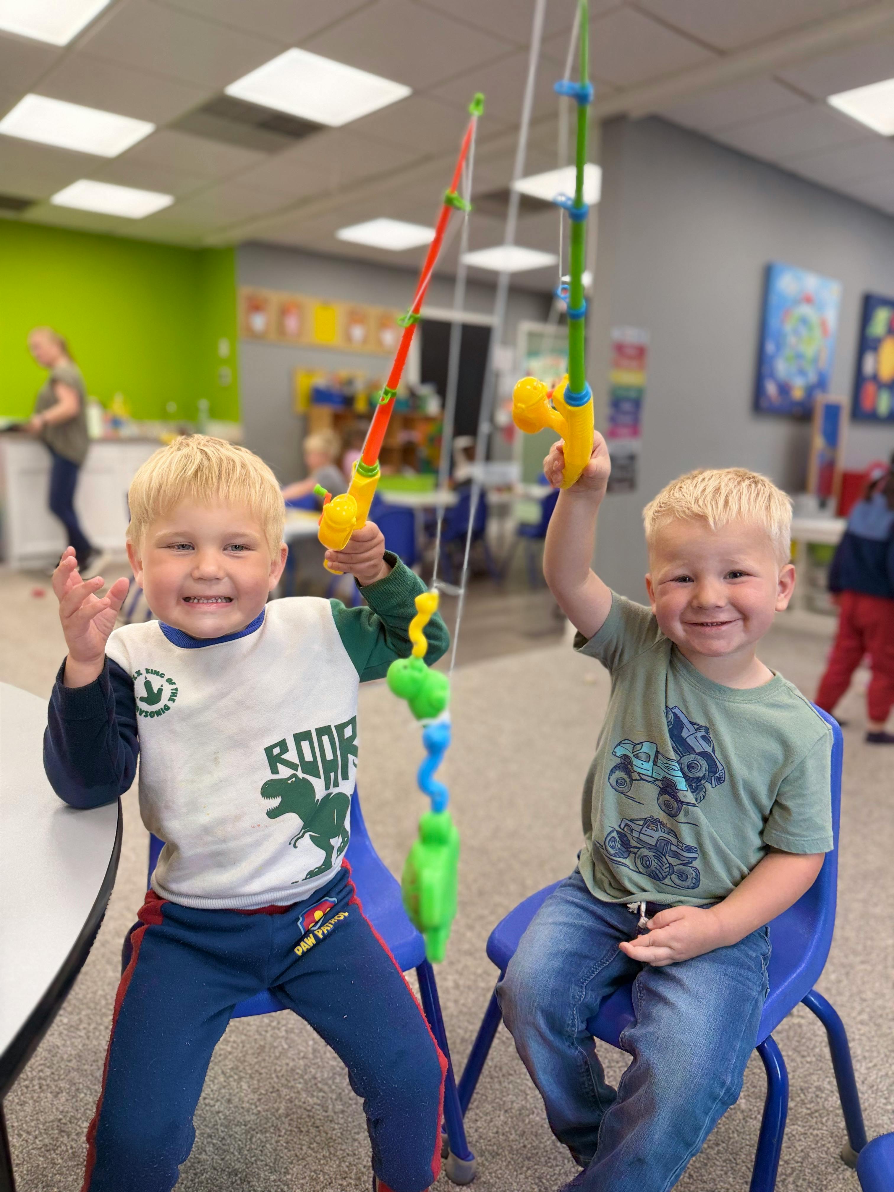 Two children sitting in chairs holding toy fishing rods in a classroom